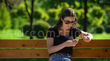 戴墨镜的女孩在阳光公园里坐在长凳上滚动智能手机，放松